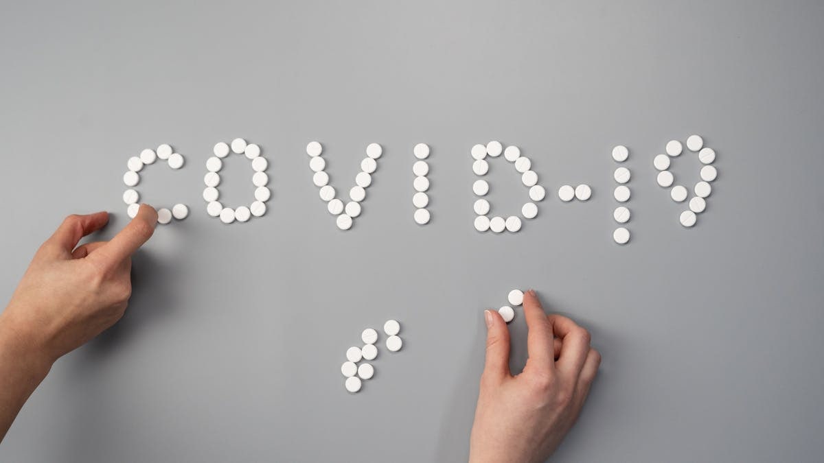Hands arranging white pills to spell 'COVID-19' on a gray surface, symbolizing medical and health themes.