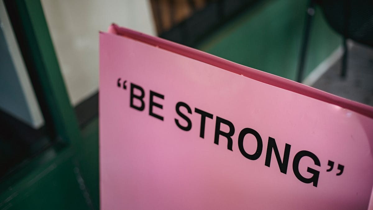 From above of pink signboard with black motivational phrase Be strong on street near green facade of building in city with blurred background